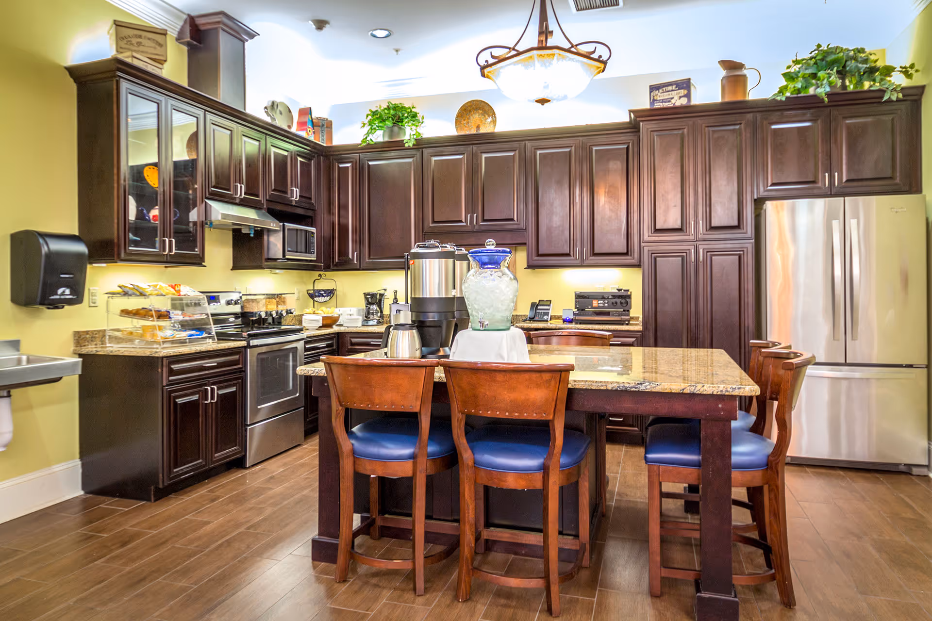 A spacious kitchen with dark wooden cabinets, stainless steel appliances including a refrigerator and oven, and a central island with a granite countertop surrounded by four wooden chairs with blue cushions. The kitchen is well-lit with a hanging light fixture and decorated with plants and kitchen items on top of the cabinets.