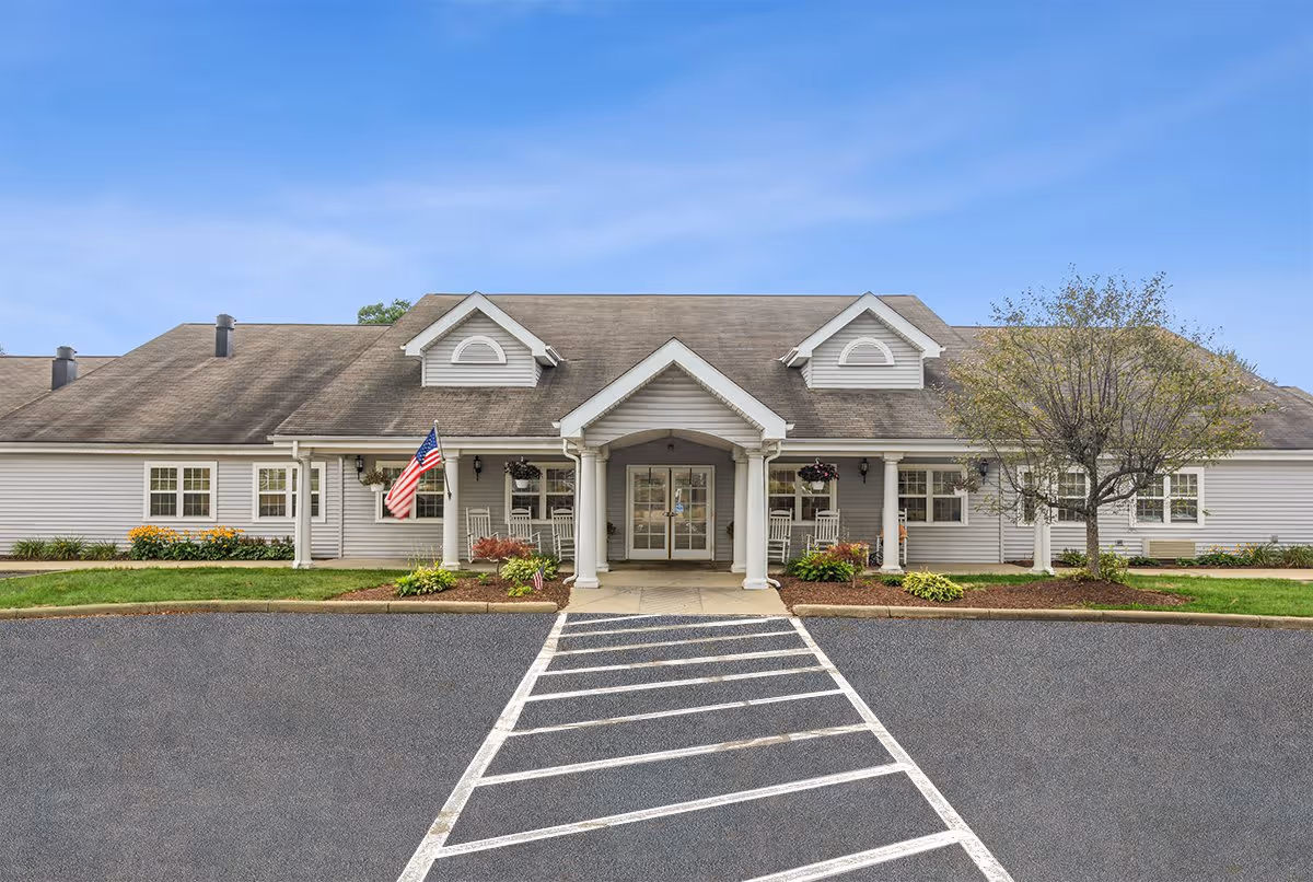 Front exterior view of a single-story senior living facility building with a covered entrance supported by white columns, an American flag, rocking chairs on the porch, landscaping with bushes and a tree, and a clear blue sky.