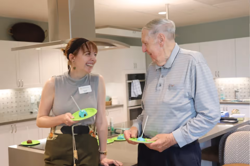A woman and an elderly man smiling and holding green plates with small desserts on them, standing and chatting in a modern kitchen with white cabinets and a kitchen island.