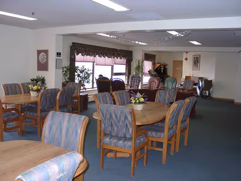 Interior view of a senior living facility common area with multiple round wooden tables surrounded by cushioned chairs with striped upholstery. There are large windows with valances letting in natural light, several potted plants, and additional seating including armchairs and a piano in the background.