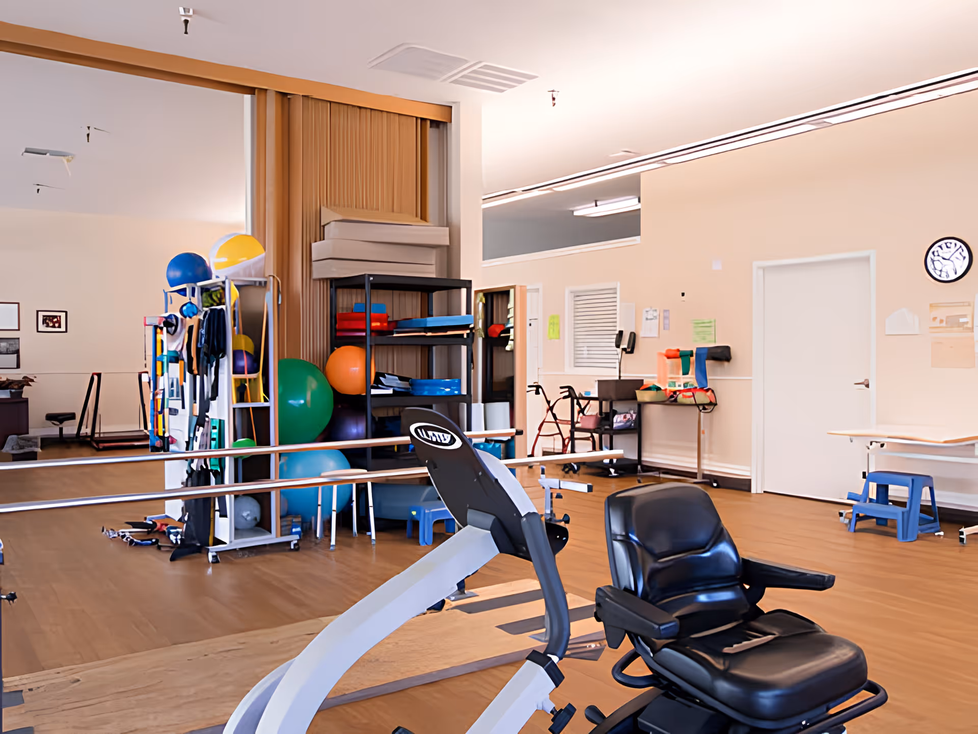 Rehabilitation gym interior with a recumbent exercise bike, parallel bars, therapy balls, and storage shelves.