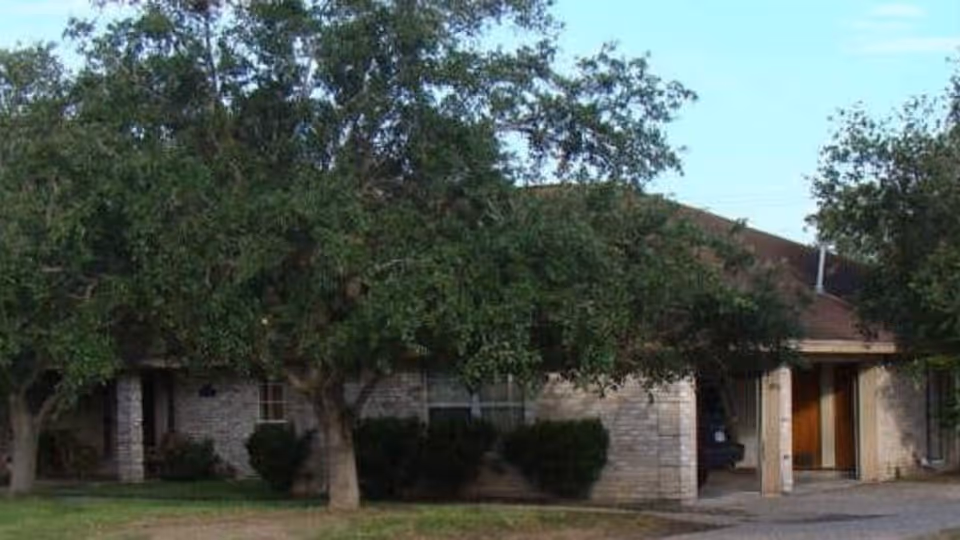 Exterior view of a single-story brick building partially obscured by large green trees and bushes, with a driveway leading to a covered entrance.