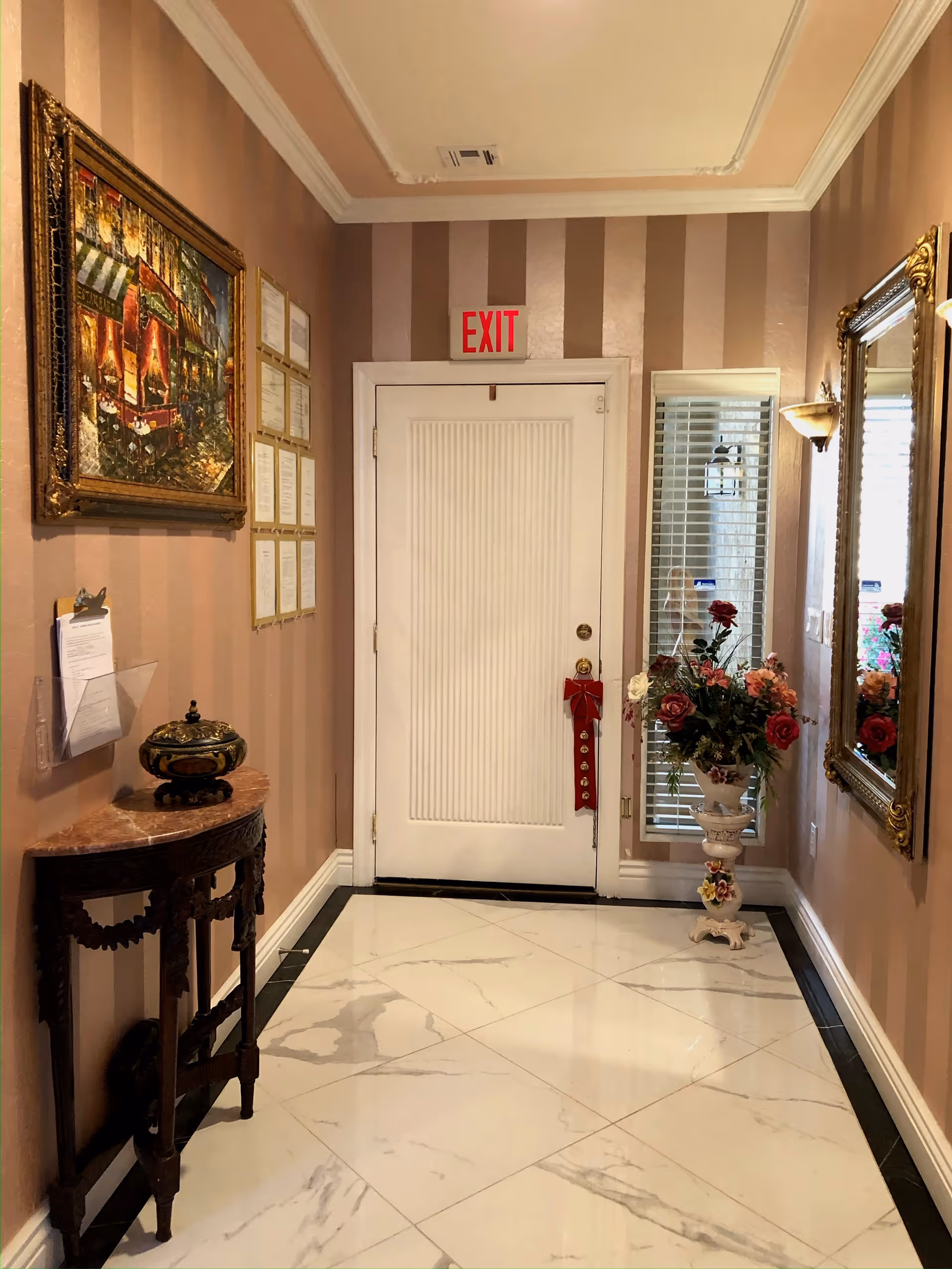 A narrow hallway with beige and brown striped walls, white marble floor tiles, and a white door with an exit sign above it. On the left wall, there is a framed painting and several certificates or documents displayed. Below them is a small wooden table with a decorative item on top. On the right side, there is a tall ornate mirror and a white pedestal holding a vase with a bouquet of colorful flowers. A wall sconce light fixture is mounted above the mirror.