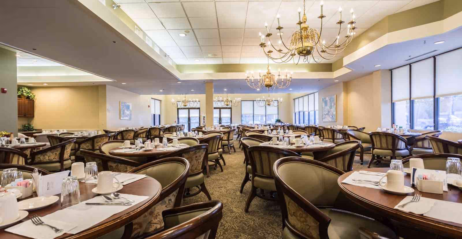 A spacious senior living dining room with multiple round tables set with white cups, glasses, plates, and silverware. The room features elegant chandeliers hanging from the ceiling, large windows with blinds allowing natural light, and comfortable upholstered chairs around each table.