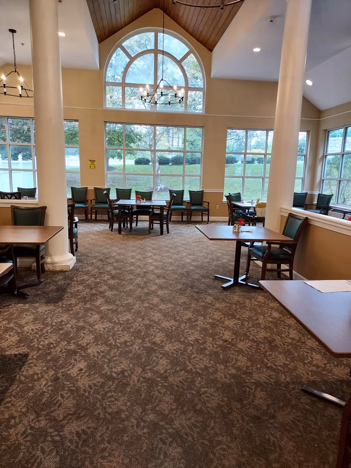 Dining room with multiple tables and chairs, tall white columns, chandeliers, and large arched windows overlooking a green lawn.