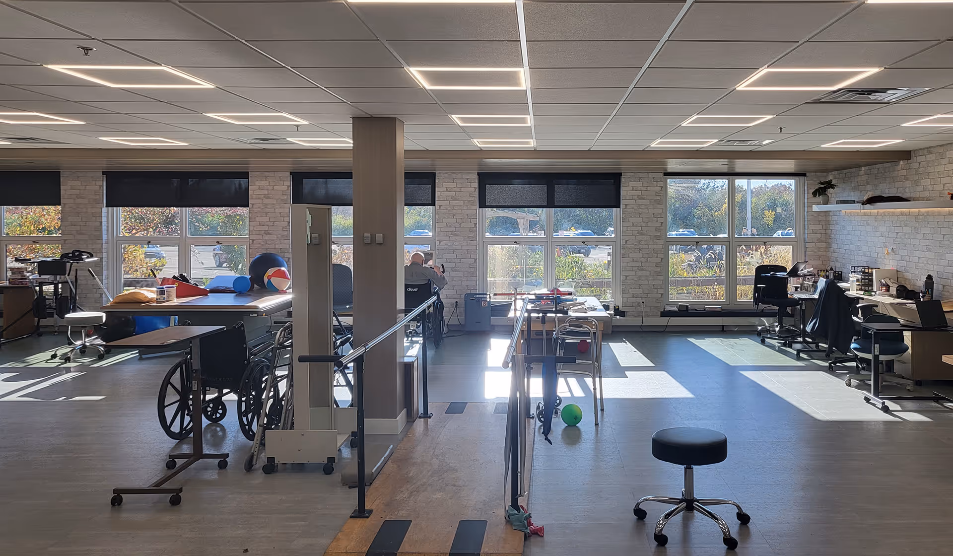 A spacious rehabilitation or physical therapy room with large windows letting in natural light. The room contains parallel bars for walking exercises, wheelchairs, therapy balls, chairs, desks with office supplies, and various therapy equipment. The walls are light-colored with a brick pattern, and the floor is a smooth surface suitable for mobility exercises.