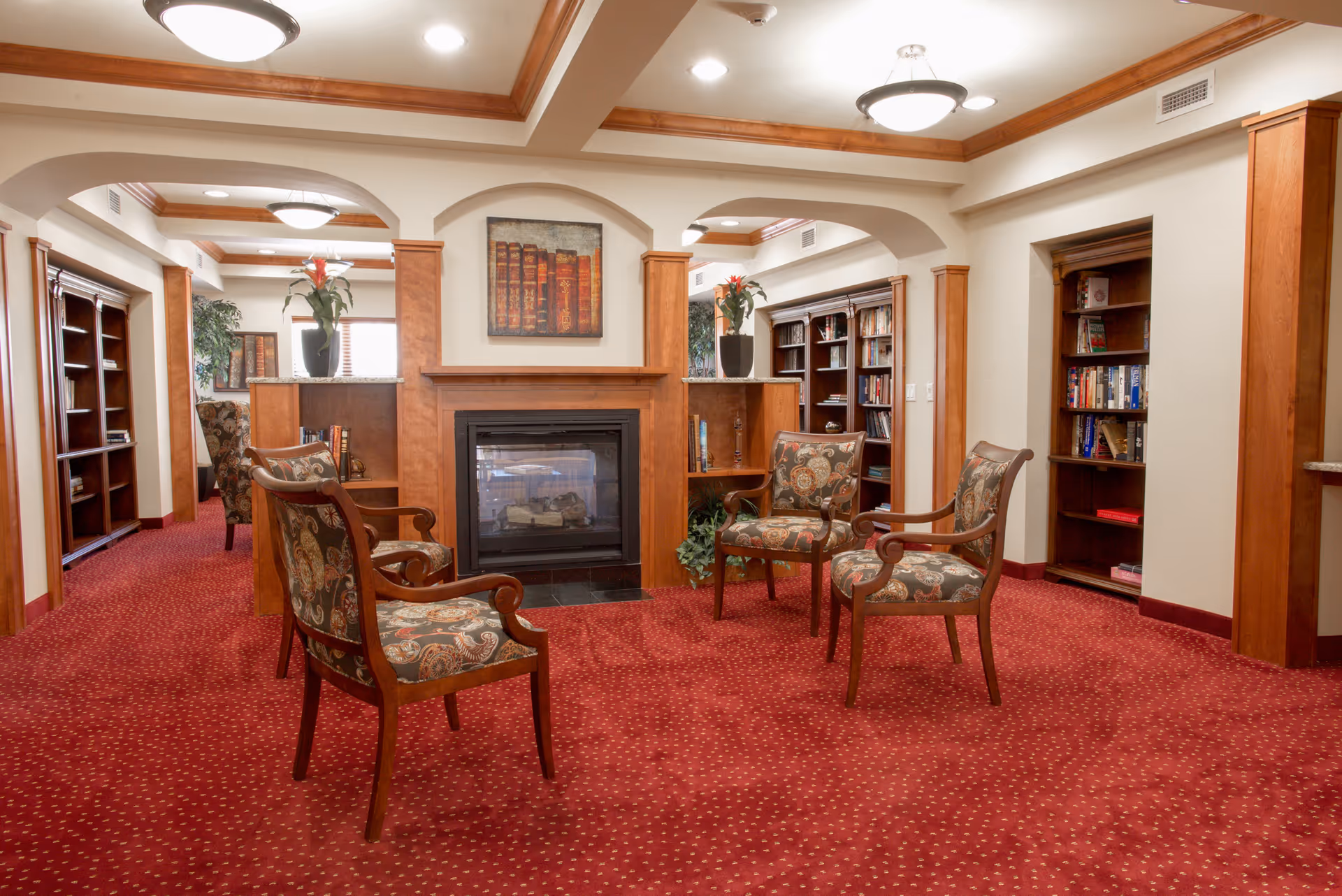 A cozy common area with four patterned armchairs arranged around a wooden fireplace. The room features red carpet with a dotted pattern, wooden bookshelves filled with books, and decorative plants on top of wooden partitions. The ceiling has recessed lighting and wooden crown molding.