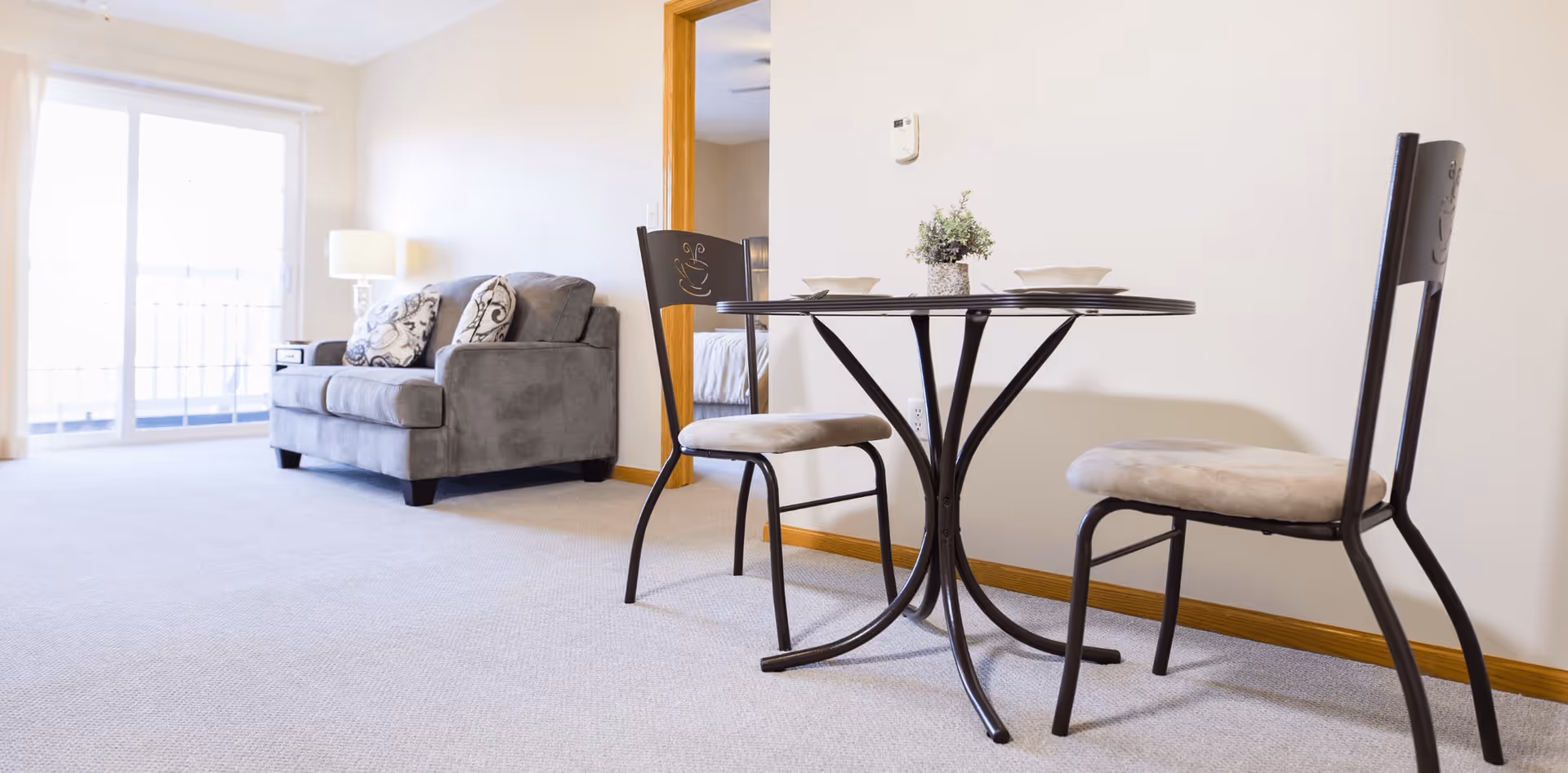 Small living area with a round bistro table and two chairs in front of a gray loveseat near a sliding glass door.