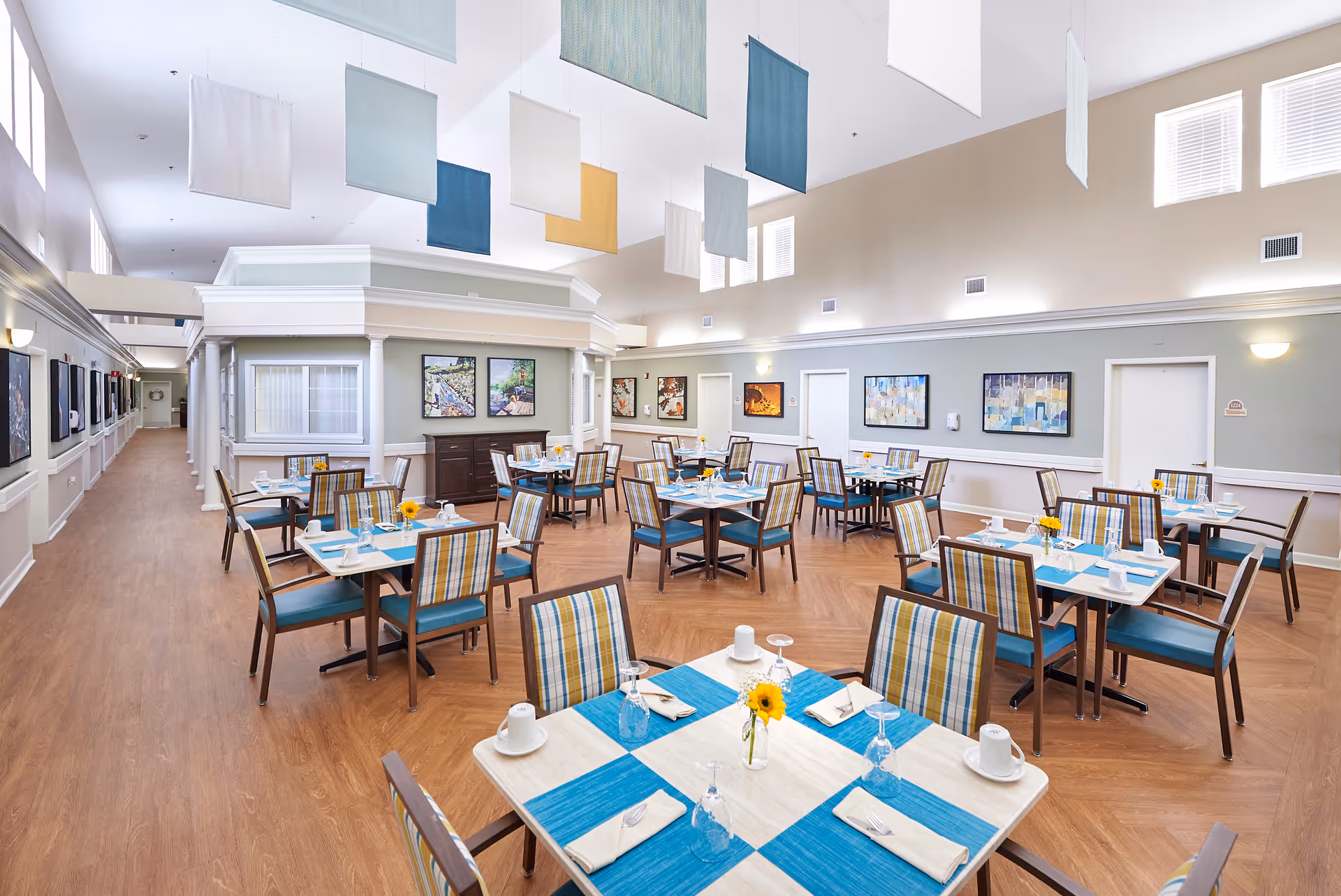 Spacious dining room with neatly set tables, striped chairs, blue table runners and colorful hanging fabric panels.