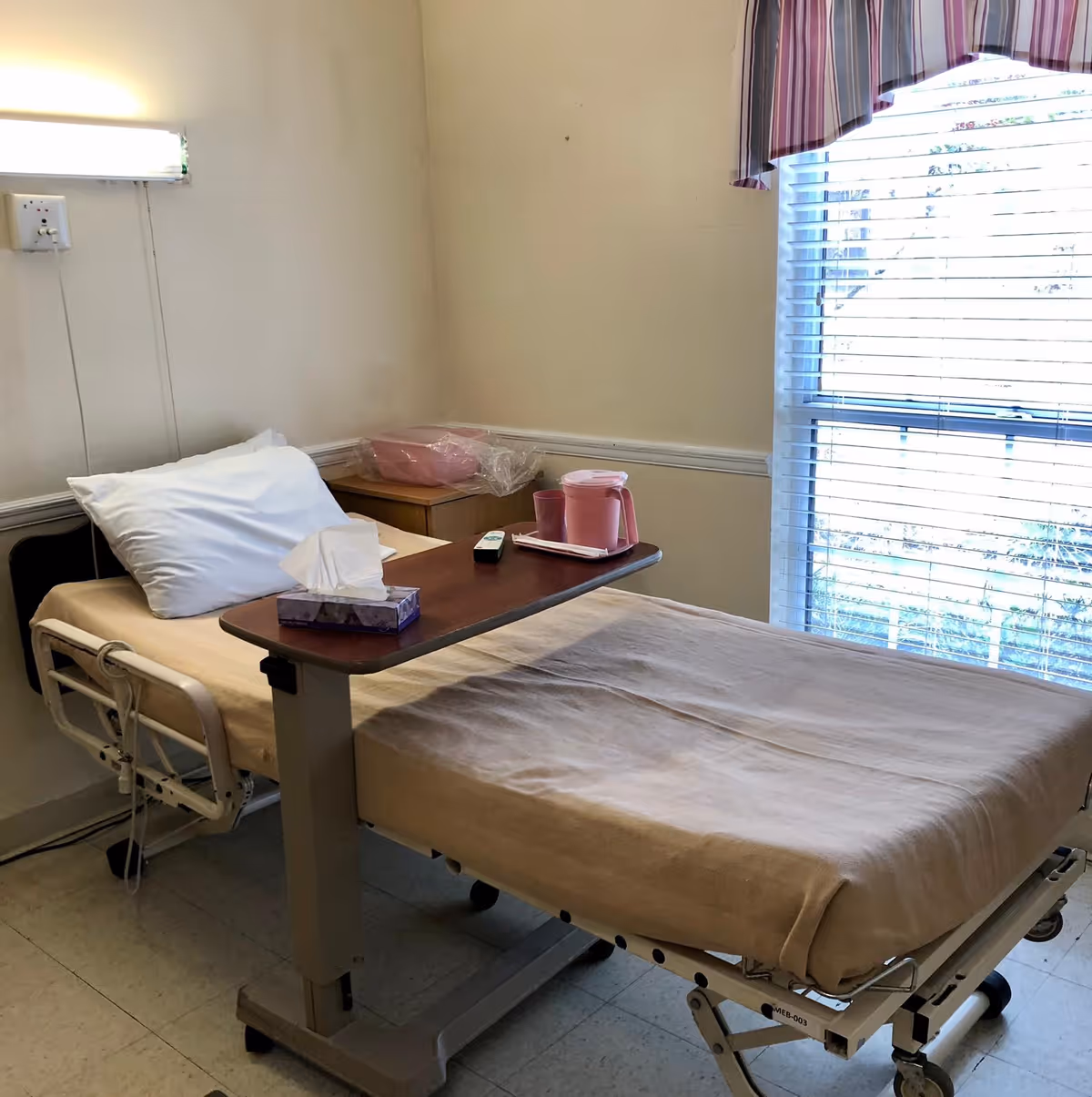 A hospital-style bed with beige bedding and a white pillow in a room. A wooden overbed table holds a tissue box, a remote control, and a pink pitcher with a cup. There is a window with blinds and a striped valance letting in natural light. A small wooden nightstand is next to the bed with a pink basin on top.