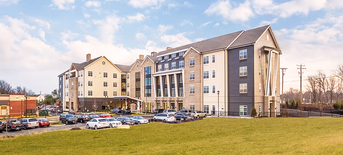 Exterior view of a large multi-story senior living facility building named Tribute at The Glen, with a parking lot filled with cars in front and a grassy area in the foreground under a partly cloudy sky.