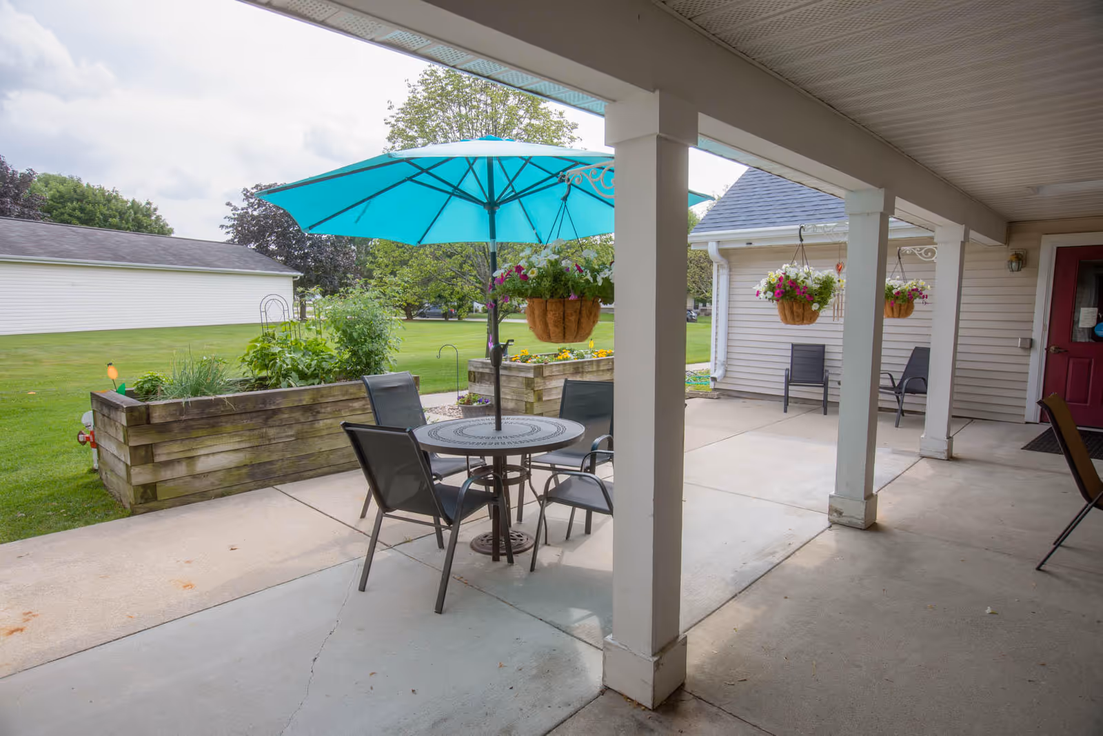 Outdoor patio area with a round metal table and four chairs under a large turquoise umbrella. There are hanging flower baskets attached to the covered porch, raised garden beds with plants, and a grassy lawn with trees in the background.