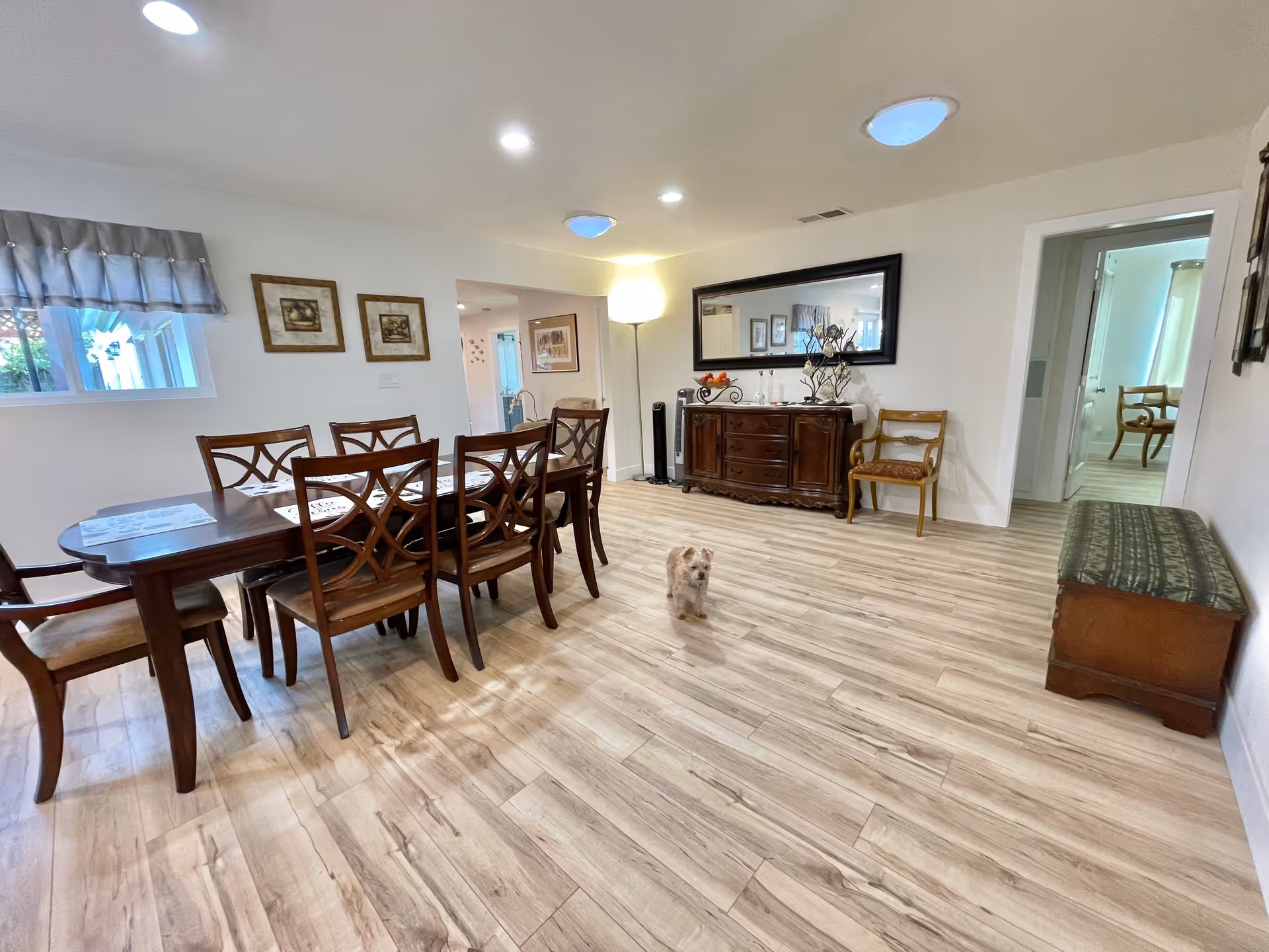 A spacious dining room with a wooden dining table and eight matching chairs. The room has light wood flooring, white walls, and a large mirror above a wooden sideboard. There are framed pictures on the walls, a floor lamp in the corner, and a small dog standing on the floor. A window with a valance lets in natural light, and there is a doorway leading to another room with more chairs visible.