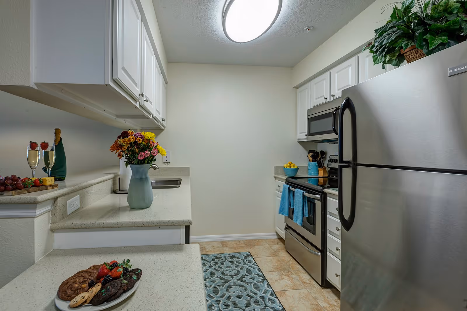 A modern kitchen with white cabinets, a stainless steel refrigerator, stove, and microwave. The countertops have a vase of colorful flowers, a plate of cookies, and a tray with two glasses of champagne and grapes. There is a blue patterned rug on the tiled floor and a potted plant on top of the refrigerator.