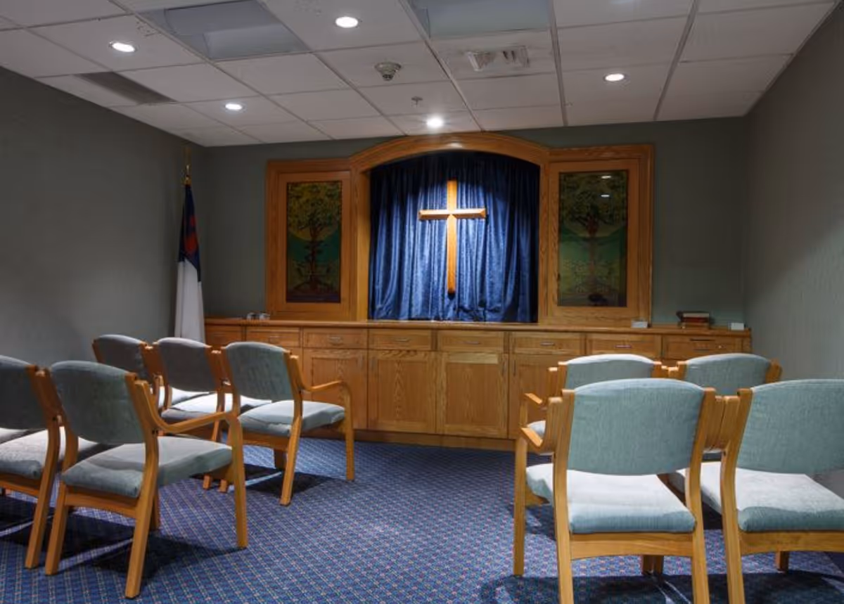 A small chapel room with a wooden cross mounted on a blue curtain backdrop, flanked by two stained glass panels depicting trees. Several wooden chairs with light blue cushions are arranged facing the cross. The room has a carpeted floor and a ceiling with recessed lighting.