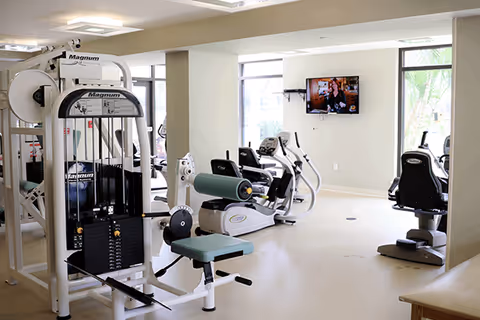 Bright exercise room with weight and cardio machines and a wall-mounted TV.