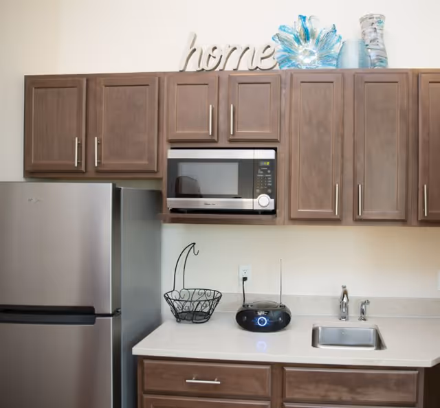 Kitchenette with stainless steel refrigerator, microwave, sink, brown cabinets and a decorative "home" sign on top of the cabinets.