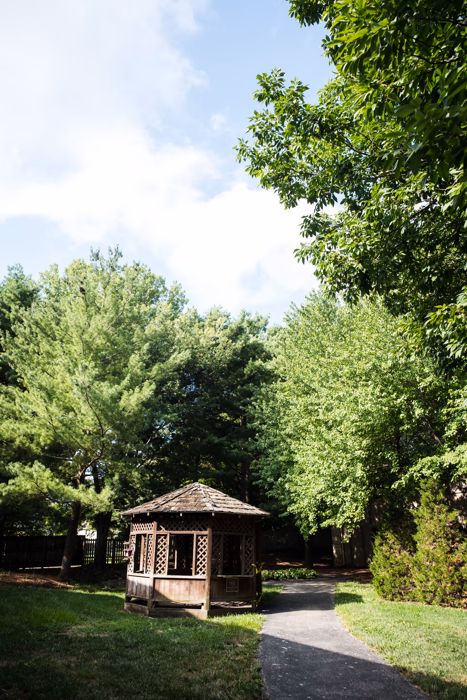 A wooden gazebo surrounded by lush green trees and grass with a paved pathway leading to it under a partly cloudy sky.
