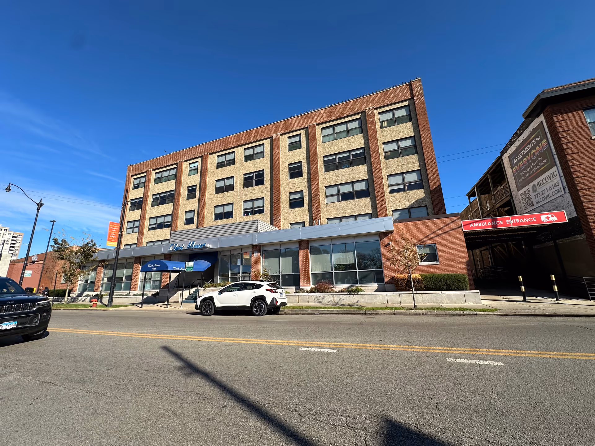 Four-story brick and concrete building labeled Clark Manor with awnings, parked cars, and a street in front under a clear blue sky.