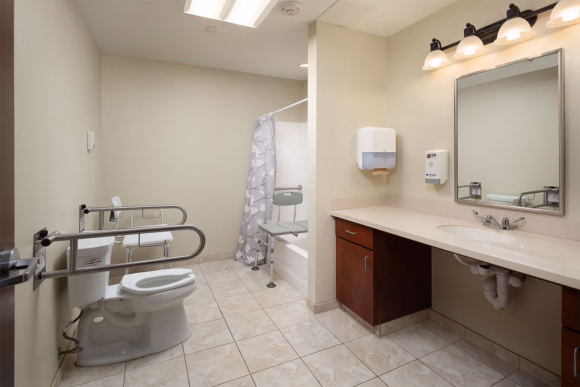 A spacious, accessible bathroom with beige tiled floor and walls. It features a toilet with safety rails, a shower with a floral-patterned curtain and a shower chair, a long countertop with an under-mount sink, a large mirror, and wall-mounted soap and paper towel dispensers. The lighting includes ceiling lights and a three-bulb fixture above the mirror.