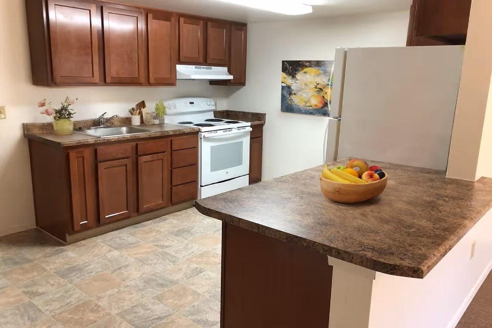 A kitchen with brown wooden cabinets, a white stove with an overhead vent, a stainless steel sink, and a refrigerator. There is a countertop with a bowl of assorted fruits including bananas, apples, and oranges. A colorful painting hangs on the wall near the refrigerator.
