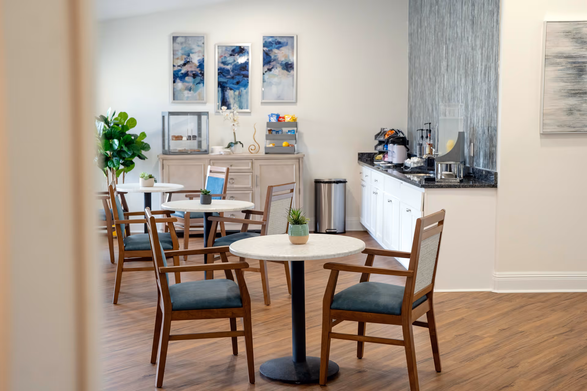 A bright and clean dining area with round tables and wooden chairs with blue cushions. There are small potted plants on the tables, a sideboard with snacks and decorations, and a counter with a coffee machine and other refreshments. The room has wooden flooring and light-colored walls adorned with abstract artwork.
