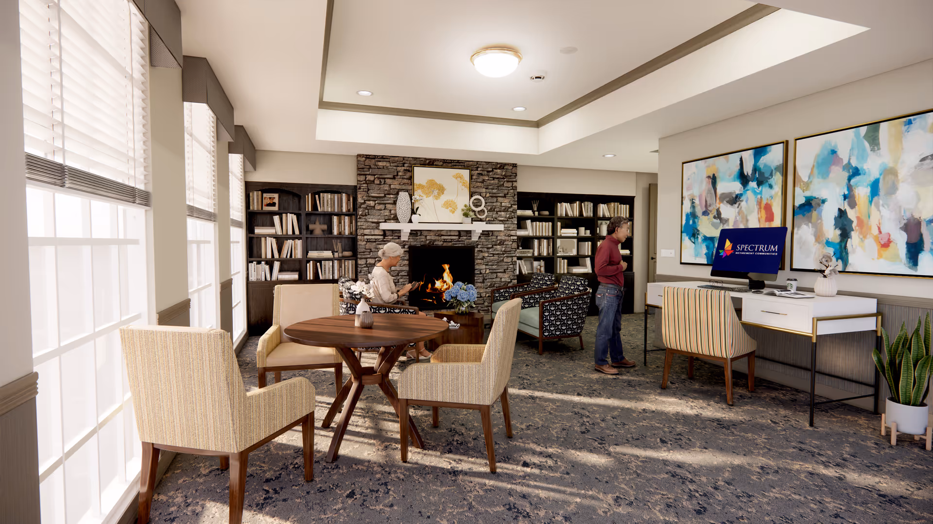 A cozy senior living common area with large windows letting in natural light. The room features a stone fireplace with a fire burning, bookshelves filled with books on either side, and comfortable seating including armchairs and a round wooden table. Two seniors are present, one seated near the fireplace and another standing near a desk with a computer displaying the Spectrum Retirement Communities logo. The walls are decorated with colorful abstract paintings and there are plants adding greenery to the space.