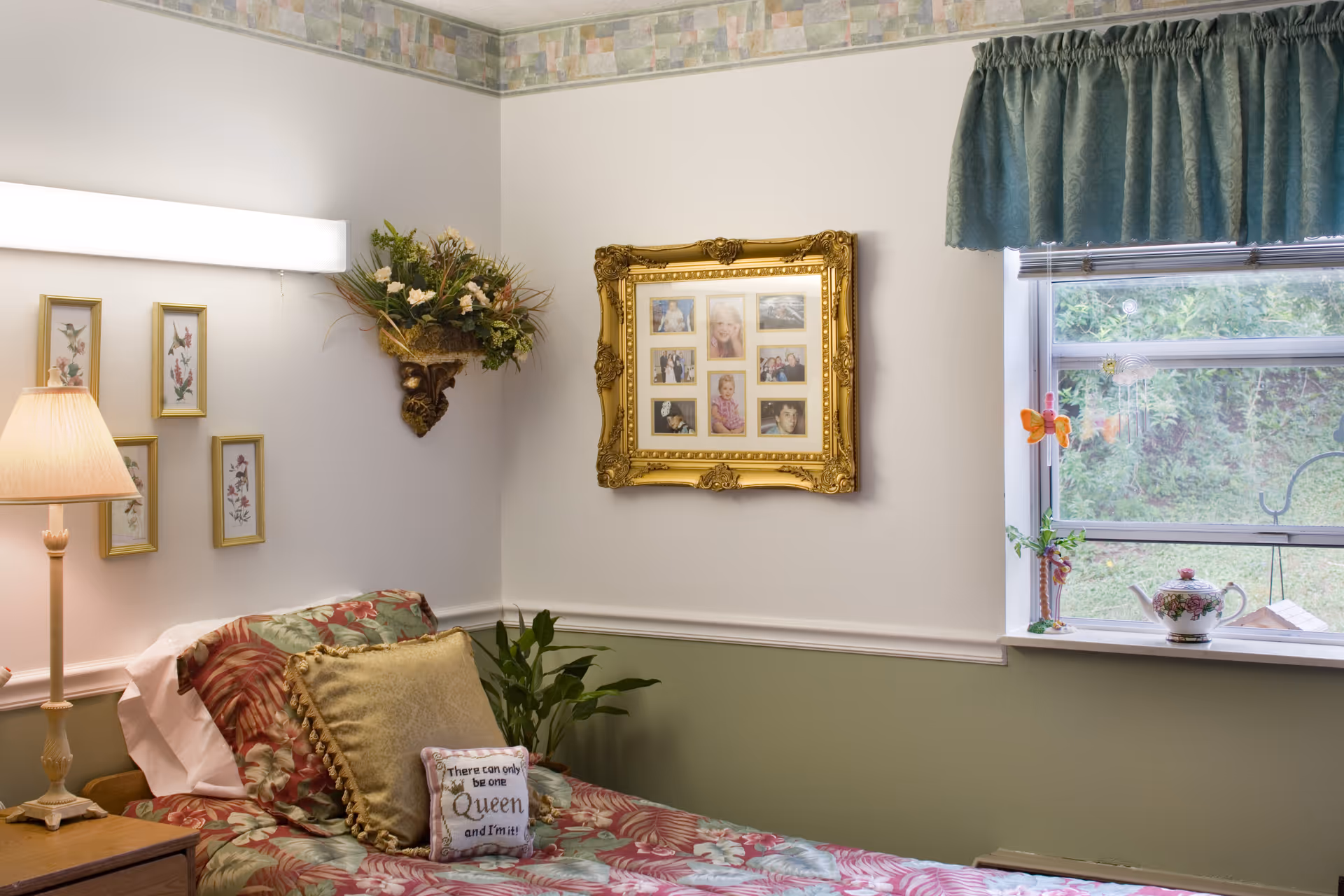 Single-bed bedroom with floral bedding, decorative pillows, a lamp, framed family photos on the wall, and a window with plants and a teapot on the sill.