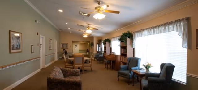 A long, well-lit common area in a senior living facility with multiple seating arrangements including armchairs and tables. The room features ceiling fans with lights, large windows with sheer curtains, framed artwork on the walls, and plants on shelves and tables, creating a cozy and inviting atmosphere.