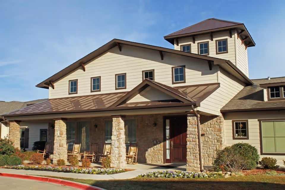 Exterior view of a senior living facility building with stone pillars supporting a covered porch. Several wooden rocking chairs are placed on the porch. The building has beige siding, multiple windows, and a brown roof with a small tower-like structure on top. The landscaping includes bushes and flower beds along the walkway.
