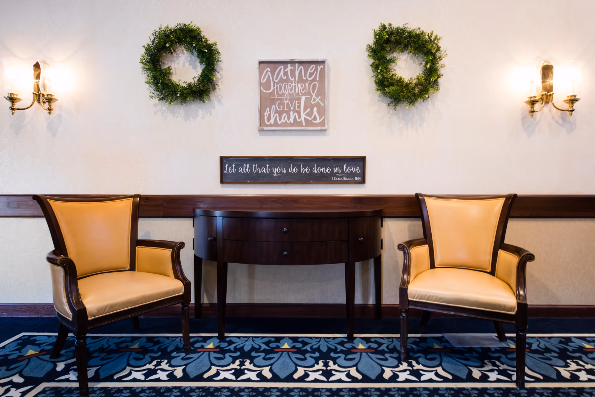 Two yellow cushioned armchairs with wooden frames are placed on either side of a dark wooden console table against a beige wall. Above the table, there are two green wreaths and two wall plaques with inspirational quotes. Two wall sconces with candles provide warm lighting on each side.