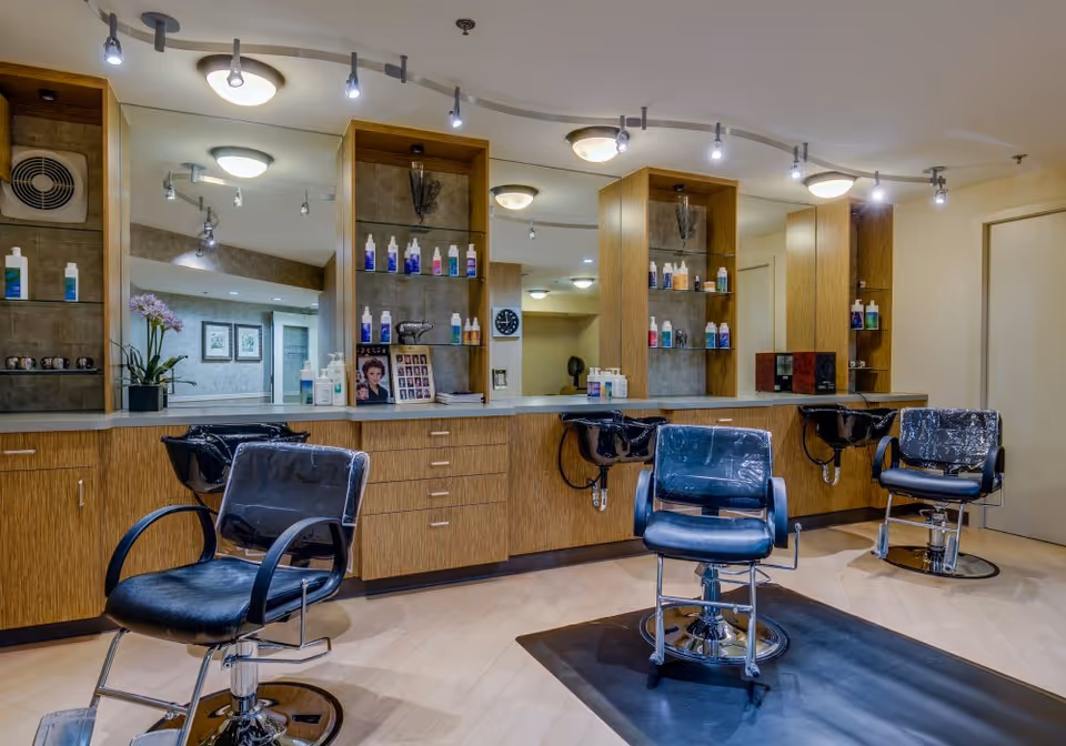 Interior view of a salon area with three black salon chairs in front of a long counter with mirrors and shelves holding various hair care products. The space is well-lit with ceiling lights and has a clean, organized appearance.