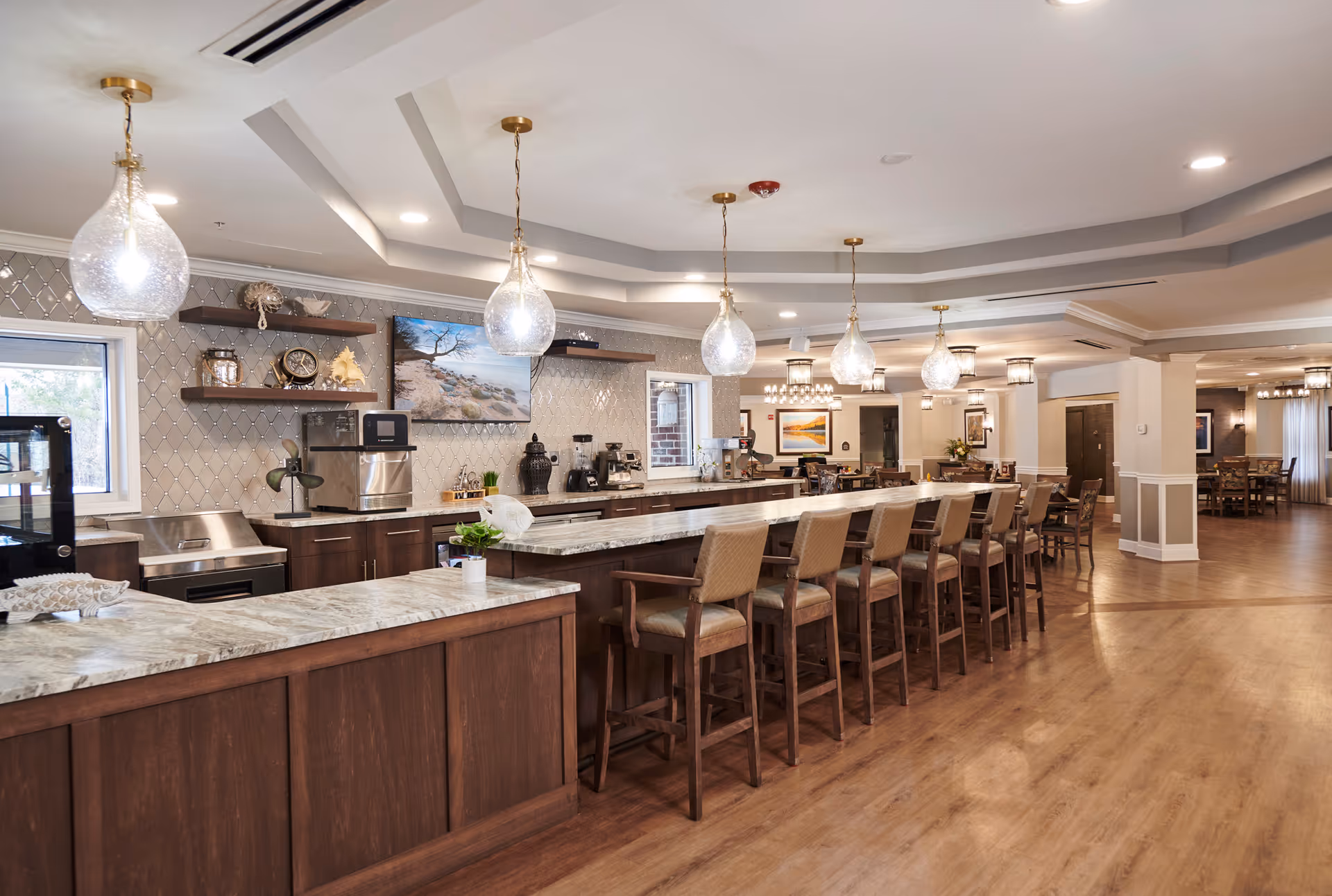A spacious and modern kitchen and dining area in a senior living facility. The kitchen features a long marble countertop island with multiple high chairs, pendant lights hanging from the ceiling, and dark wood cabinetry. The background shows additional dining tables and chairs, decorative lighting fixtures, and framed artwork on the walls.