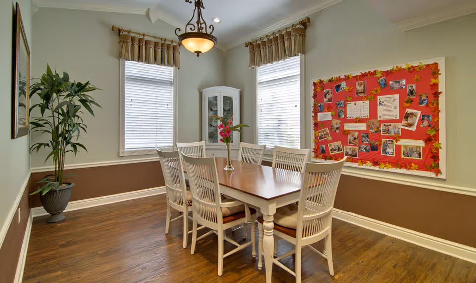 A dining room with a wooden table and six white chairs. The room has two windows with blinds and valances, a potted plant in the corner, a white corner cabinet, and a bulletin board with photos and decorations on the wall. The floor is wooden, and a chandelier hangs from the ceiling.