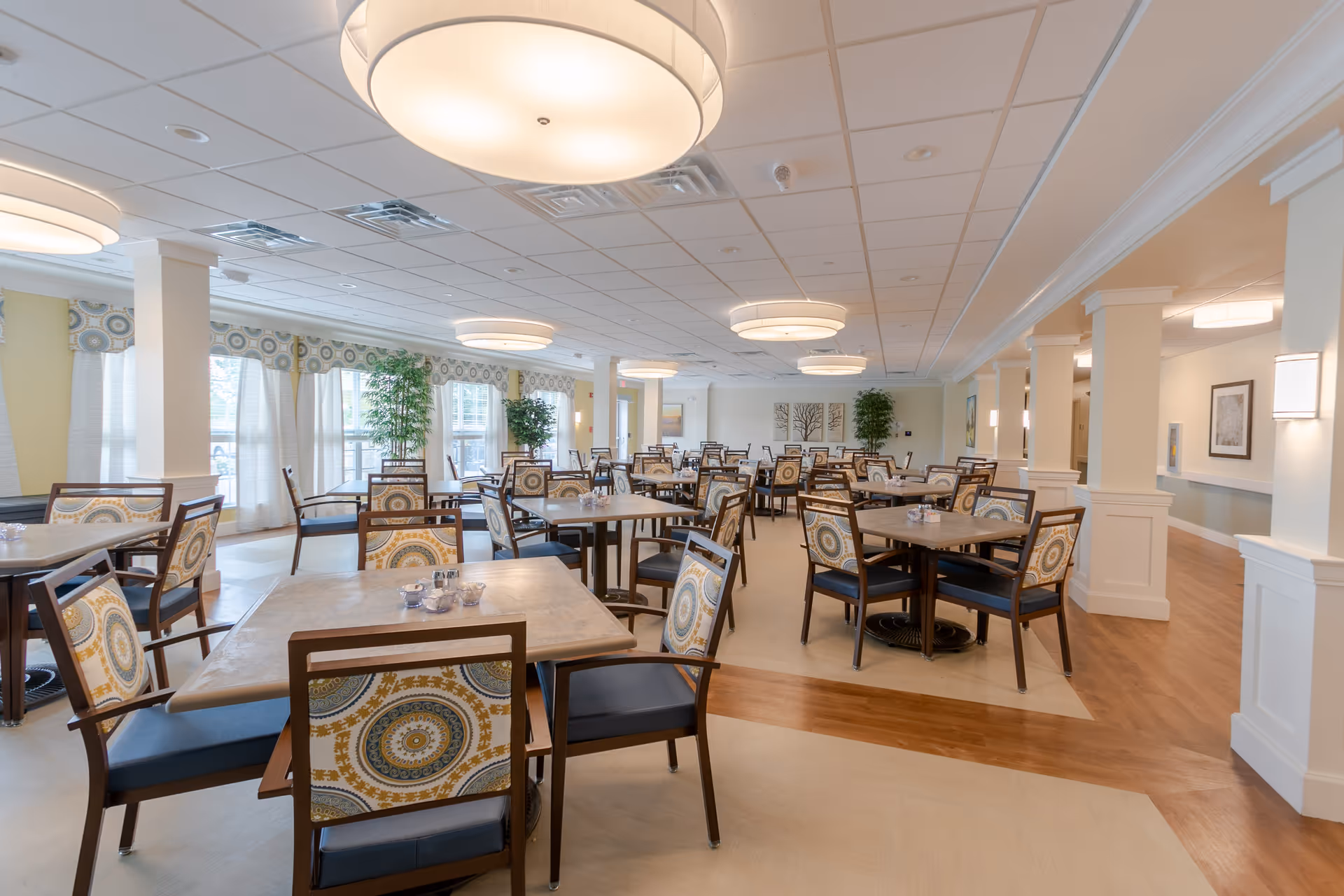 A spacious dining room in a senior living facility with multiple square tables and chairs featuring patterned upholstery. The room has large windows with curtains, several ceiling lights, and decorative plants. The floor has a combination of light-colored and wood-patterned sections.