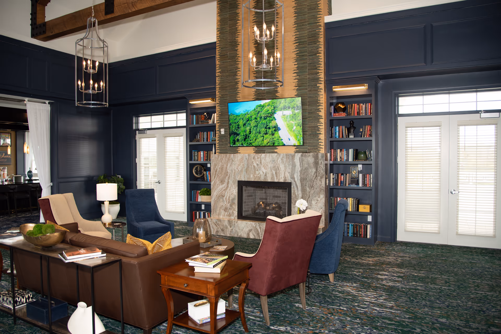 Communal living room with sofas and armchairs arranged around a fireplace and wall-mounted TV, flanked by built-in bookshelves and double doors.