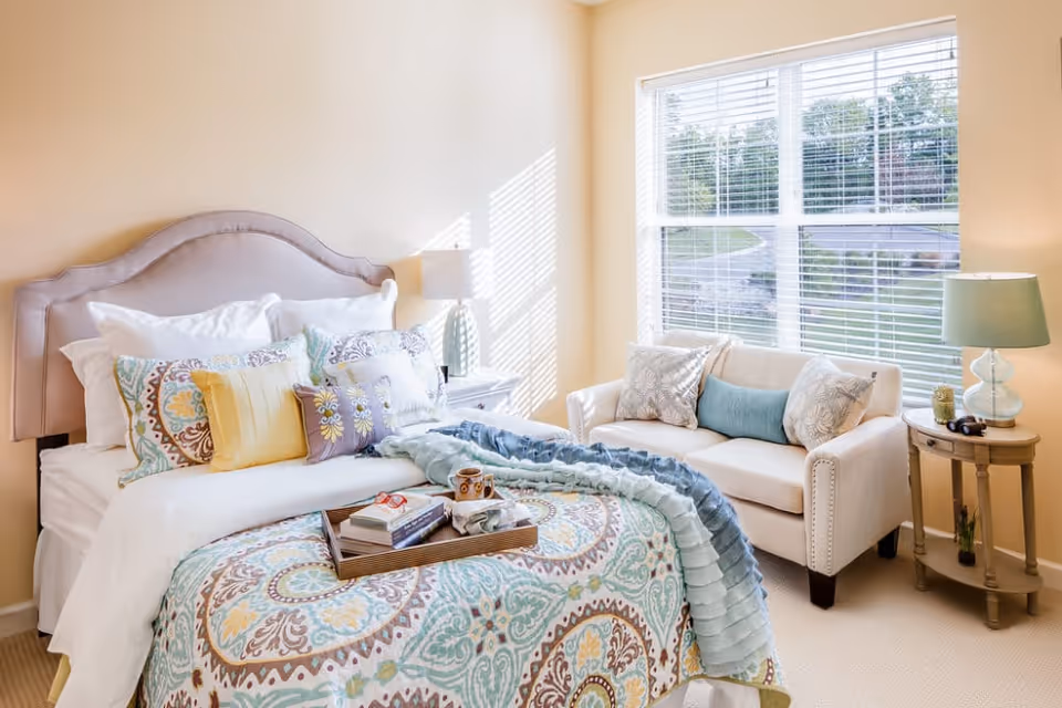 Sunlit bedroom with a decorative bed, patterned bedding and tray, and a small loveseat by a large window.