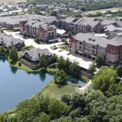 Aerial view of a senior living facility named Watercrest at Mansfield, showing multiple buildings with gray roofs, surrounded by greenery and a pond in the foreground.