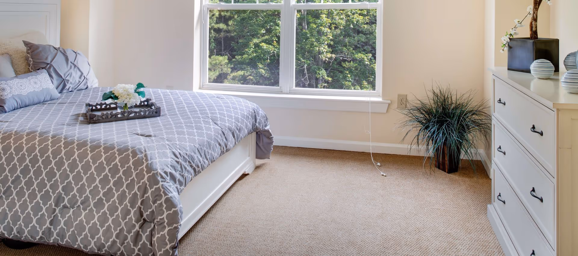 A bright bedroom with a large window showing green trees outside. The bed has a gray and white patterned comforter with several pillows and a tray with white flowers on top. There is a white dresser with decorative vases and a plant in a pot on the floor next to it. The walls are light-colored and the floor is carpeted.