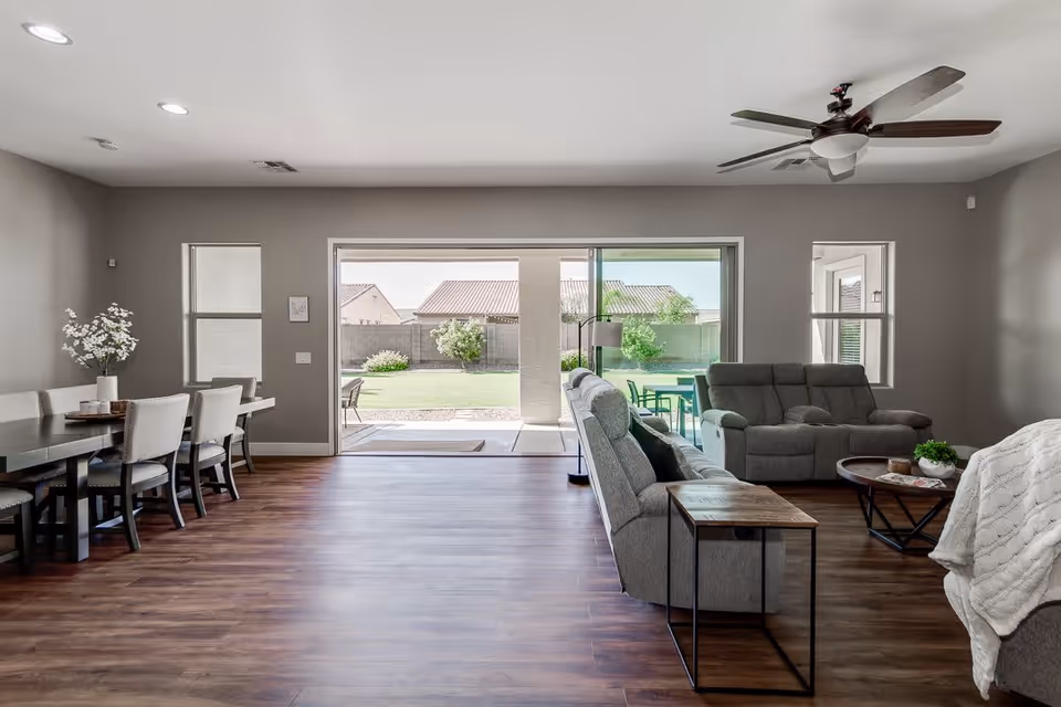 A spacious living and dining area with wooden flooring. The dining area on the left features a dark wooden table with six upholstered chairs and a vase with white flowers. The living area on the right has two gray recliner sofas, a round wooden coffee table, and a side table. Large sliding glass doors open to a backyard with green grass, bushes, and a concrete patio with outdoor furniture. The ceiling has recessed lights and a ceiling fan with dark wooden blades.