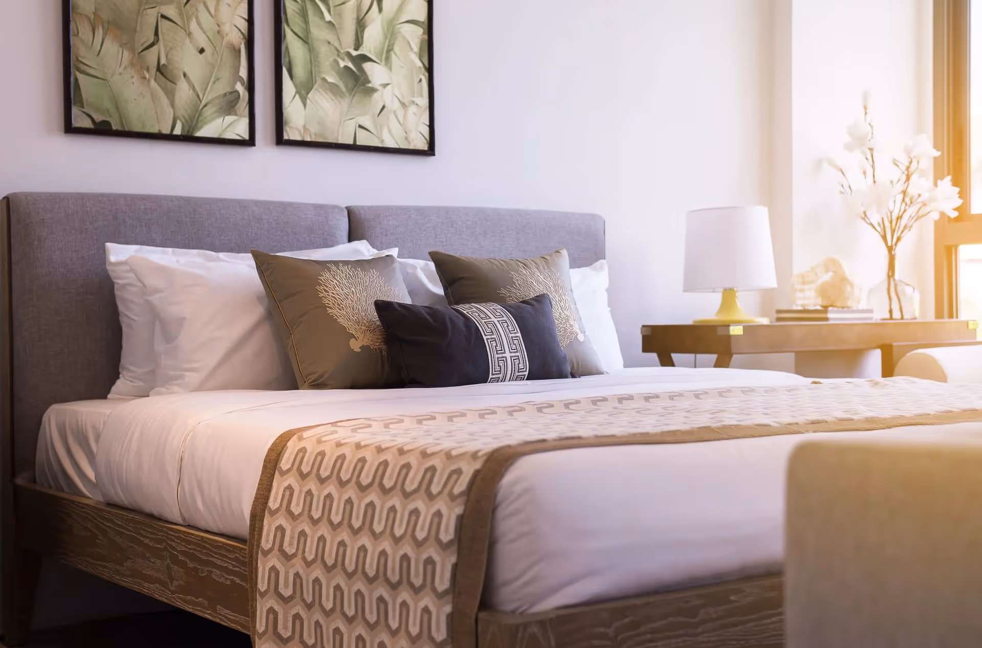 Well-lit bedroom with a made bed, decorative pillows, framed wall art, and a bedside table with a lamp.