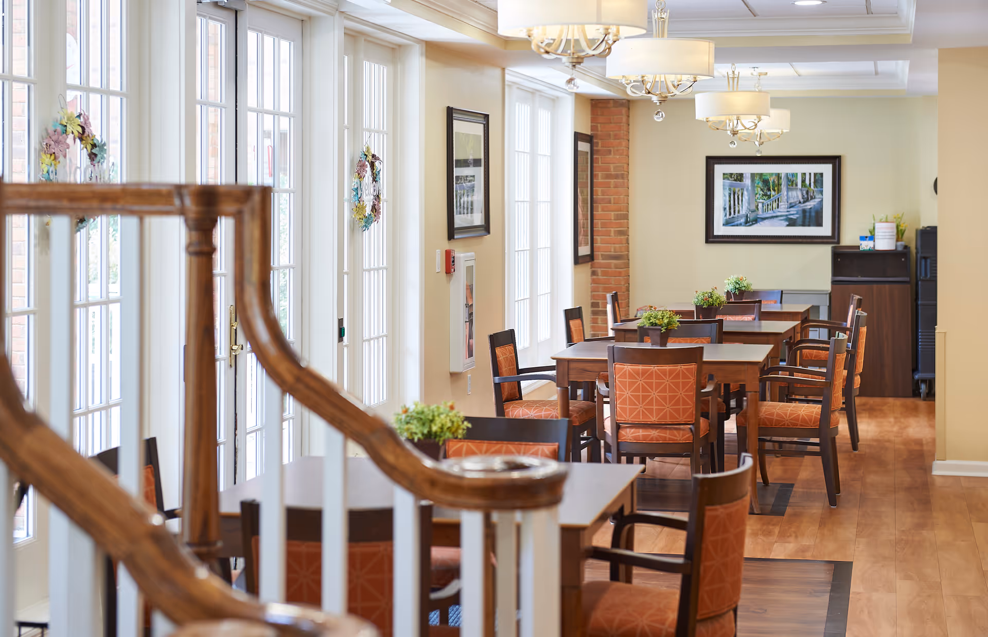 Dining room with multiple wooden tables and orange upholstered chairs, large windows along one wall, and ceiling chandeliers.