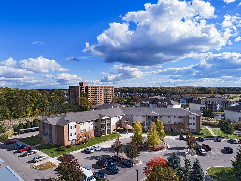 Aerial view of a senior living facility named American House Southgate, showing a large multi-story building surrounded by parking lots with cars, trees with autumn foliage, and other residential buildings in the background under a partly cloudy blue sky.