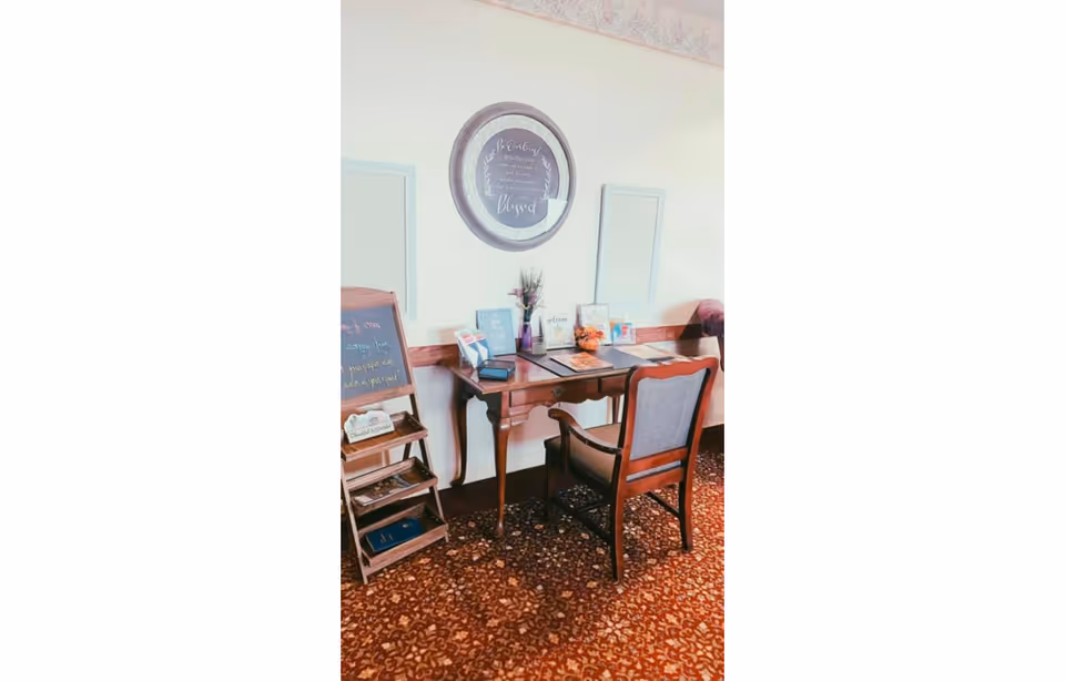 A small wooden desk with a cushioned wooden chair in front of it, placed against a light-colored wall. On the desk are various decorative items including framed pictures, a small vase with flowers, and a bowl with orange objects. Above the desk hangs a round framed sign with a quote. To the left of the desk is a small wooden stand with a chalkboard sign and some brochures or pamphlets. The floor is covered with a patterned carpet in shades of brown and red.