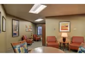 Small communal lounge with upholstered chairs, end tables, framed artwork, and a round table under fluorescent ceiling lights.