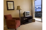 A cozy living room area with a red armchair, a floor lamp, a small TV on a dark wooden stand, a wall mirror, and a sliding glass door leading to a balcony with a view of buildings and water.
