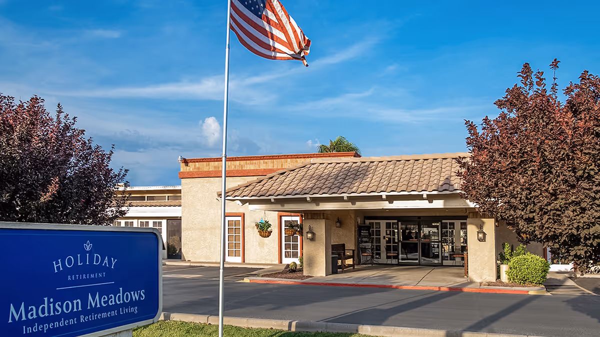 Entrance of Madison Meadows Retirement Community with a covered drop-off area, an American flag on a flagpole, and a blue sign that reads 'Holiday Retirement Madison Meadows Independent Retirement Living'. Trees with reddish leaves flank the entrance under a clear blue sky.