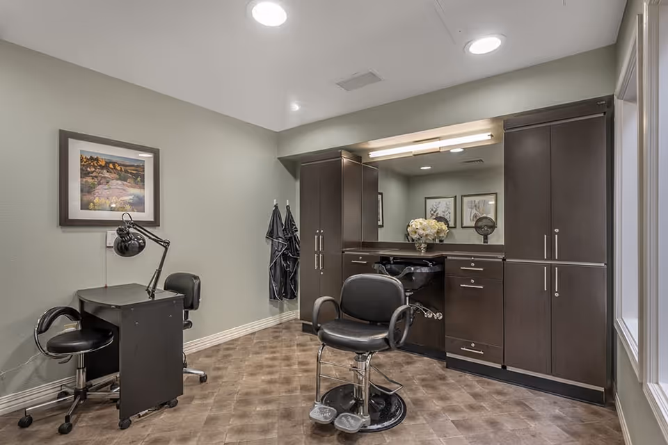 Interior view of a salon room with a black salon chair in front of a large mirror and dark wood cabinetry. There is a small black desk with a lamp and two black chairs on the left side, a framed landscape picture on the wall, and a window on the right side letting in natural light.
