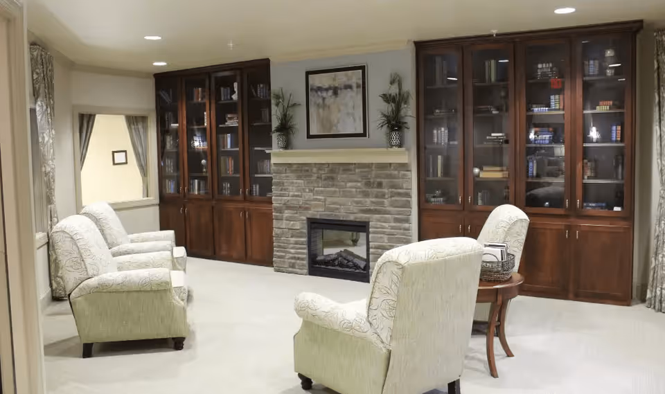 A cozy living room area with four upholstered armchairs arranged around a small wooden table. The room features a stone fireplace with a mantel decorated with two potted plants and a framed abstract painting above it. On either side of the fireplace are tall wooden cabinets with glass doors filled with books and decorative items. Light-colored curtains frame a window on the left side of the room.