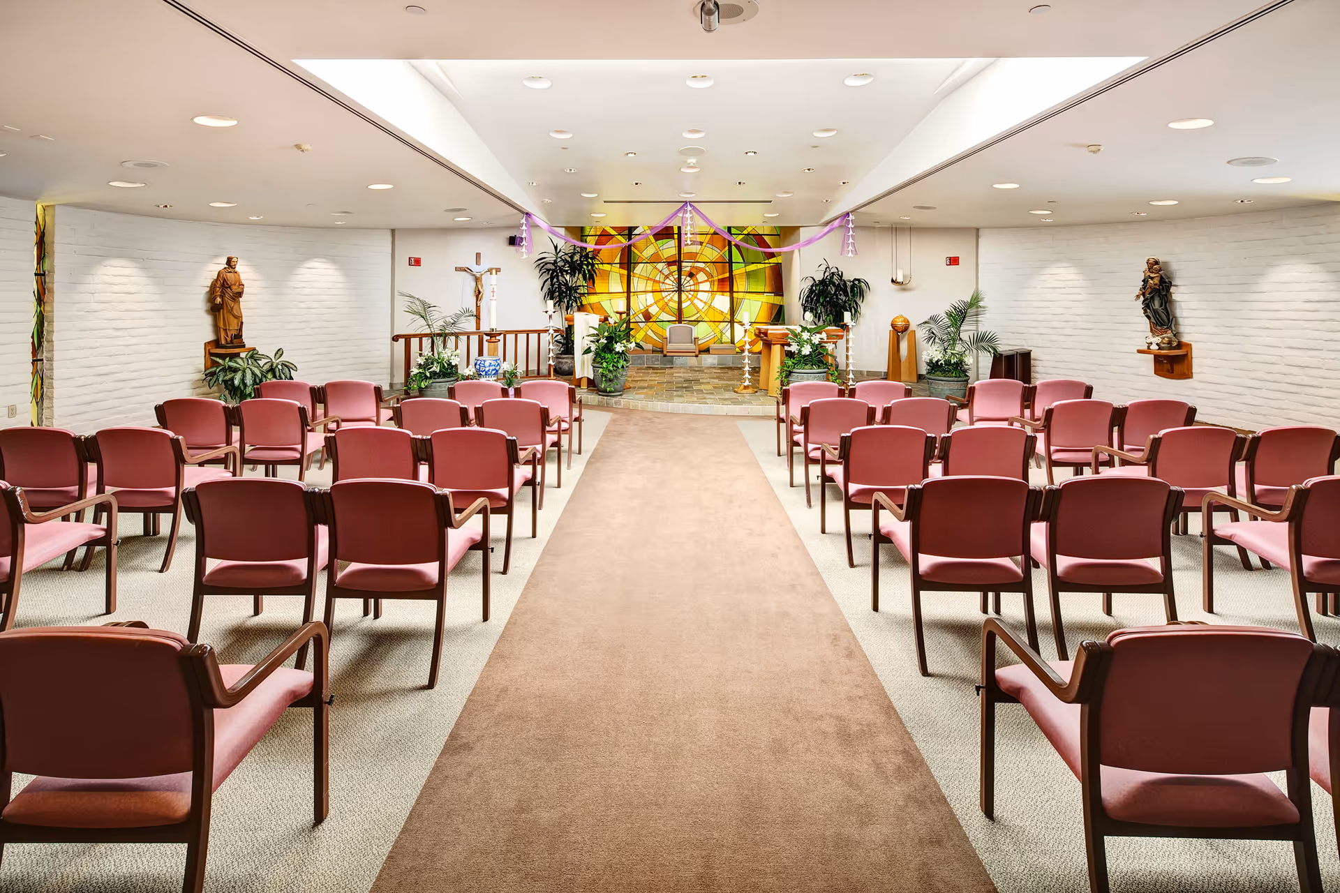 Interior view of a chapel or worship room with rows of pink cushioned chairs facing an altar area. The altar features a large colorful stained glass window, religious statues, plants, and candles. The room has white brick walls and a beige carpeted aisle leading to the altar.